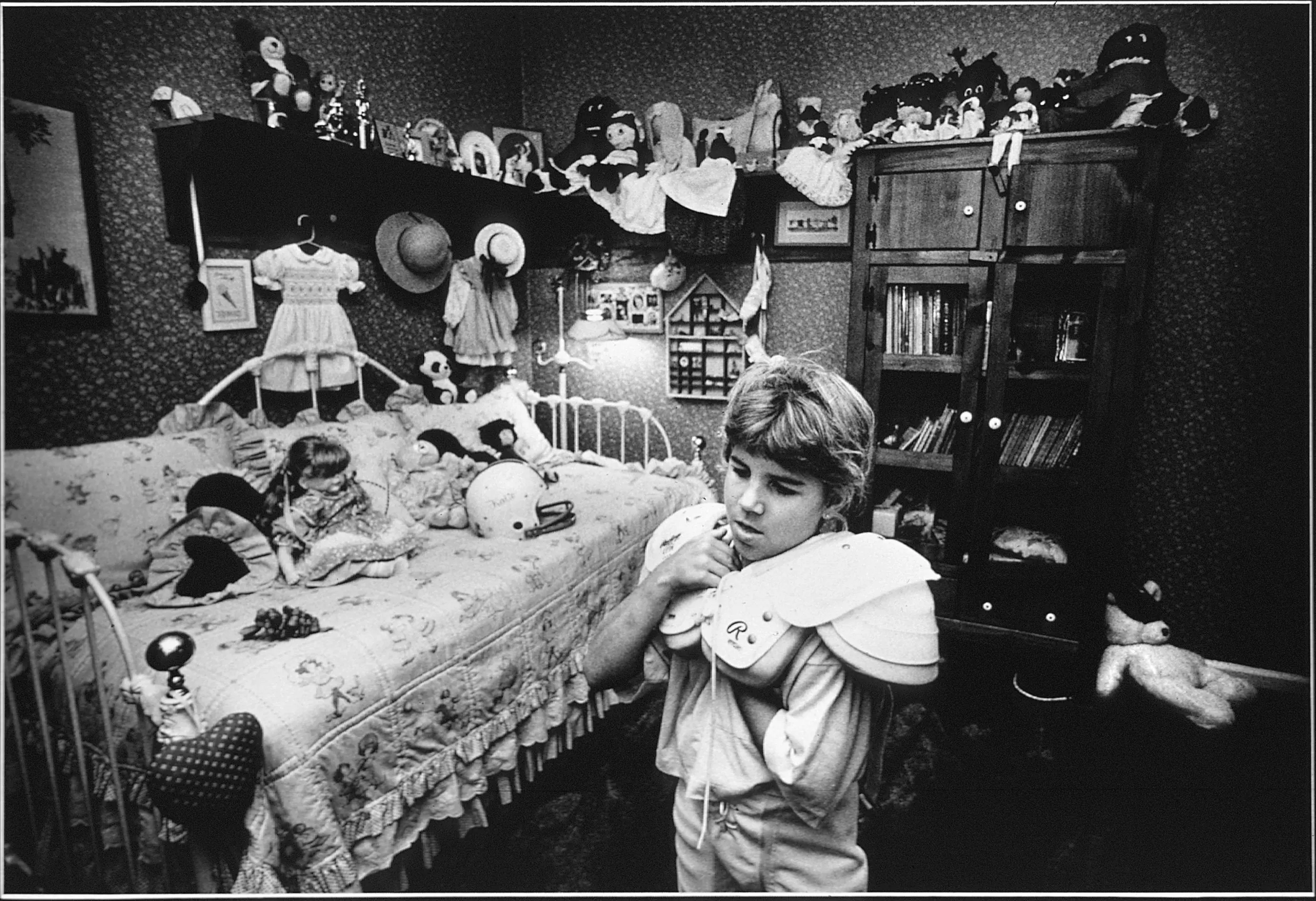 Dressing Up — Surrounded by her dolls, Katie Wood, an eight-year-old member of an all boys pee-wee football team, dons her shoulder pads in her bedroom before leaving home for practice in Baton Rouge, Louisiana. Katie is the only girl playing in an all-boys league.  (Photo by Stephan Savoia)  Published by The Baton Rouge Louisiana Sunday Advocate Magazine on December 3, 1989