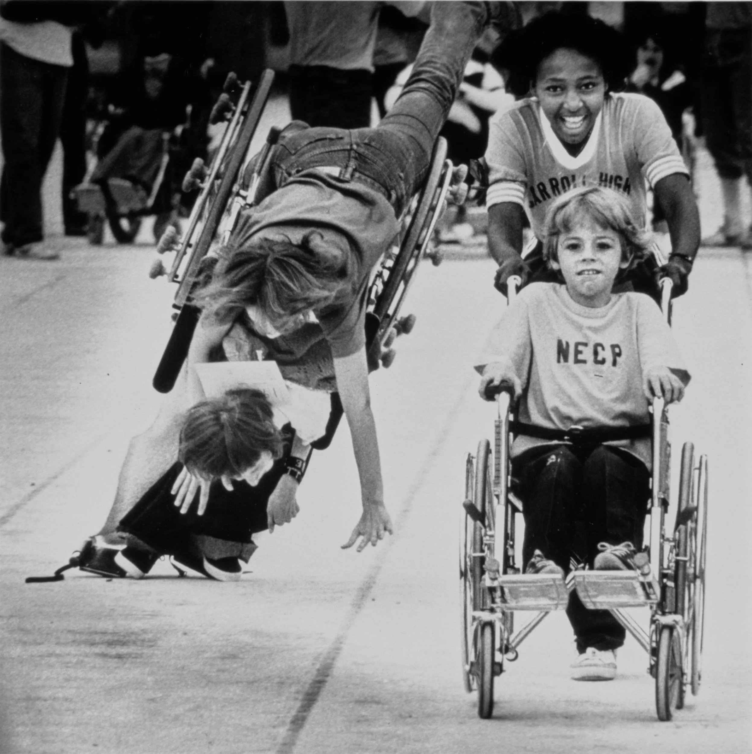 Wheelchair Crash — Chris Green, 9, at right, of the Northeast Louisiana Cerebral Palsy Flyers, is pushed by his runner, Yolanda Davis, as they head toward a first place win in the 25 meter wheelchair dash at the Region 9-A Special Olympics in Monroe, Louisiana on Saturday April 4, 1982. Also of the Flyers team, runner Elise Stark, 19, topples head-over-wheelchair as she competes with her sister Judy Stark. Judy fell out of her chair before the Starks righted themselves and completed the race. Except for a few scratches neither sister was injured and both were proud and happy to have competed.  (Photo by Stephan Savoia)  Published by The Monroe Louisiana News-Star-World