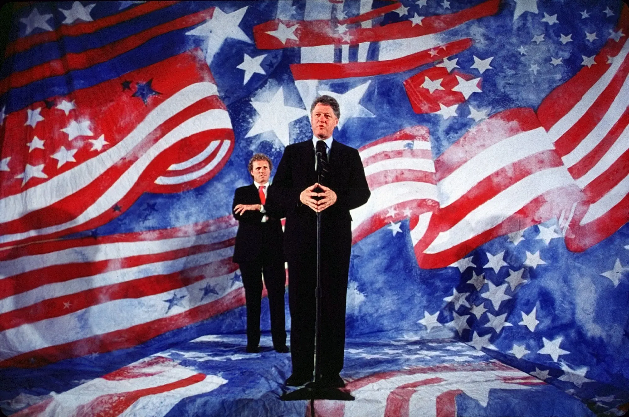Star Spangled Stage — Democratic presidential hopeful, Arkansas Governor Bill Clinton, addresses the media in Boston, Massachusetts while speaking via satellite to Pennsylvania voters on the night of his Pennsylvania presidential primary victory as U.S. Representative Joseph Kennedy, Democrat Massachusetts, looks on Tuesday, April 28, 1992. Typically a presidential candidate will address their primary voters in a state hosting its primary; Clinton, however, chose to use his time to tap Massachusetts donors for campaign contributions. Clinton won the Pennsylvania primary by 56.5 percent, more than twice the number of votes as his nearest competitor.  (AP Photo/Stephan Savoia)  © The Associated Press