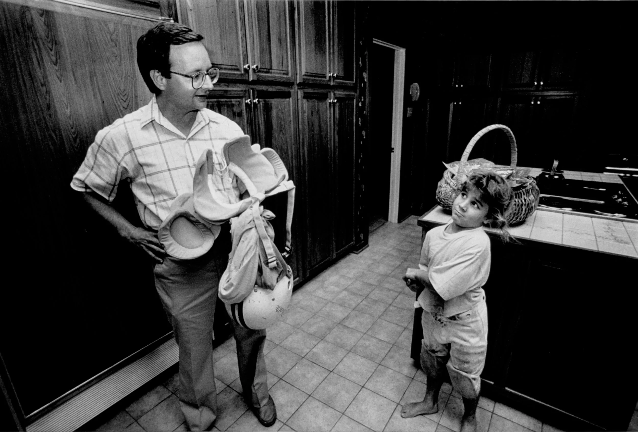 Post Game Locker Room — Standing in the kitchen of their Baton Rouge, Louisiana home eight year-old Katie Woods, removes her dirty football uniform handing her helmet, shoulder pads and jersey to her father, Dr. Danny Woods, after her final game of the season. Katie scored a touchdown during the game which ended in a tie.  (Photo by Stephan Savoia)  Published by The Baton Rouge Louisiana Sunday Advocate Magazine on December 3, 1989
